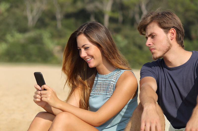 eifersüchtiger Partner schaut Freundin am Strand auf ihr Handy